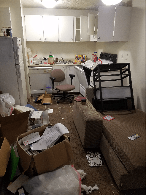Messy kitchen and living area, with clutter on the floor, cabinets, and appliances.