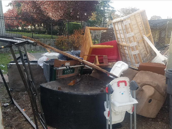 Pile of discarded furniture and debris on the side of a road, including a high chair, chairs, and wood.